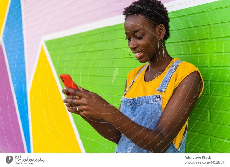 Smiling black woman texting on smartphone near colorful wall using watch bright surfing style positive text message internet female mobile online browsing