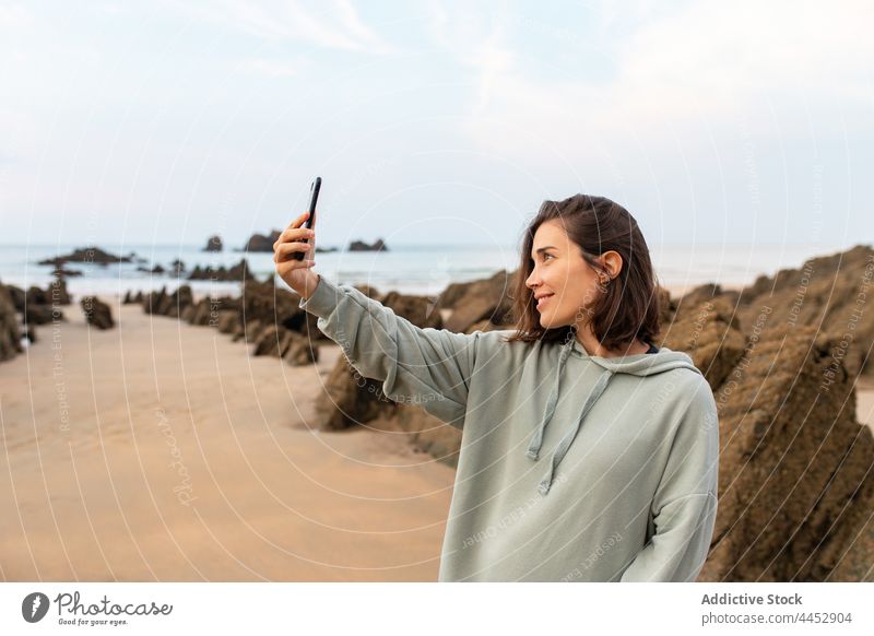 Smiling woman taking selfie on smartphone on rocky beach self portrait seacoast memory moment sky free time using gadget device cellphone watching pastime
