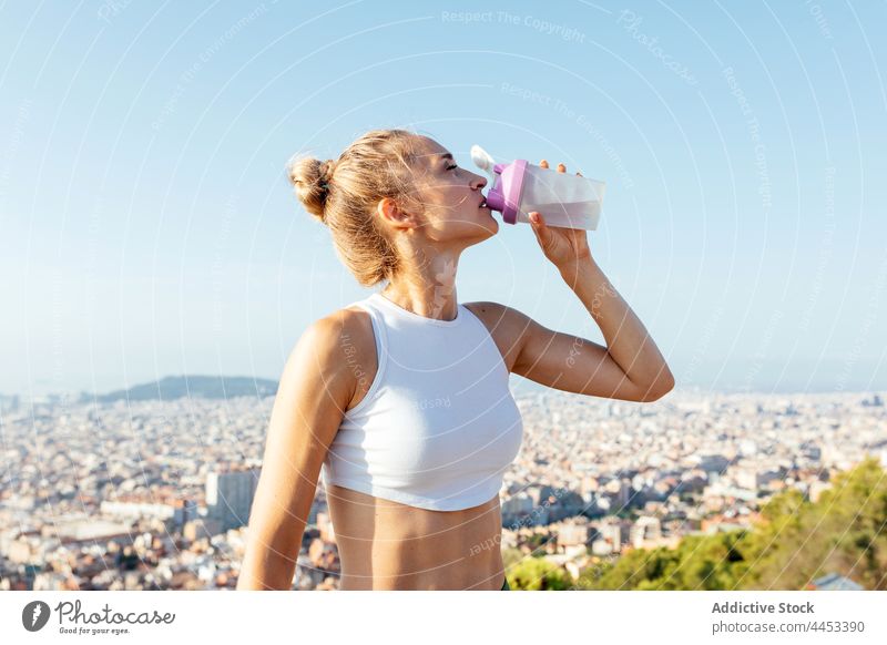 Fit sportswoman drinking water under light sky in sunny city athlete hydrate thirsty hand on hip sporty body break bottle aqua refreshment beverage