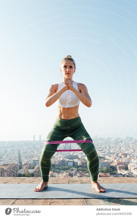 Fit sportswoman squatting with resistance band during workout on rooftop hands clasped training active warm up exercise healthy lifestyle energy vitality city