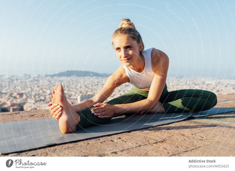 Woman stretching leg on yoga mat in town woman forward bend healthy lifestyle touch foot wellness vitality rooftop blue sky city pose practice wellbeing energy