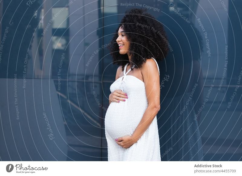 Smiling pregnant woman in long white dress happy expect posture enjoy charming appearance carefree personality female pregnancy glad curly hair afro positive