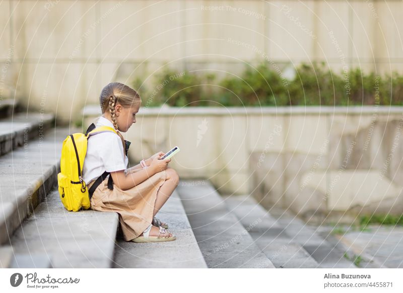 Pupil wearing school uniform and backpack outdoor. little caucasian blonde girl is playing with mobile phone. back to school Education online video chat