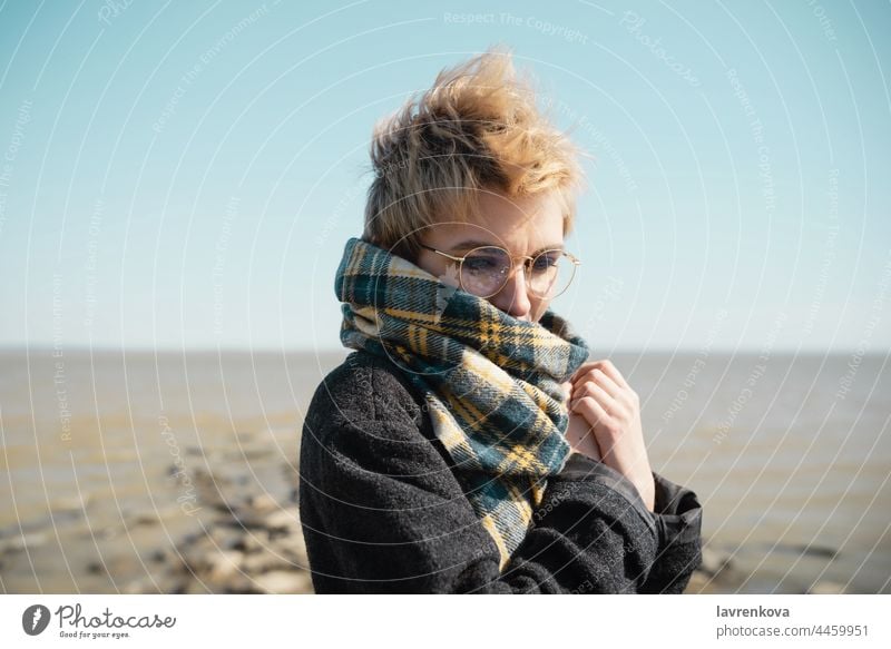 Young female in glasses warming herself on a beach alone coat autumn portrait young vacation face caucasian nature outdoors seaside sky travel water woman cold