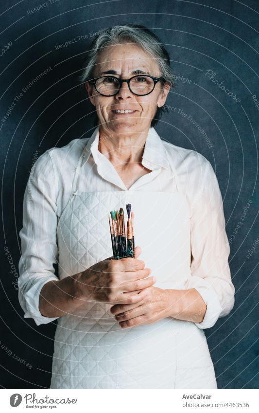Smiling older woman, proud artist, with grey hair and glasses and many paintbrushes, artistic image creativity hobby holding portrait chair colours education
