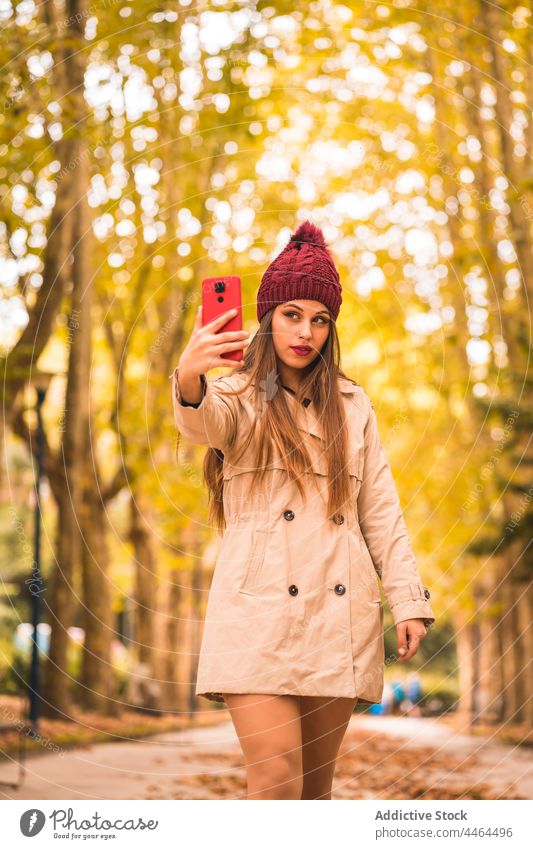 Woman taking selfie on smartphone in autumn park woman style using alley fall feminine female trench coat self portrait take photo tree mobile device gadget hat