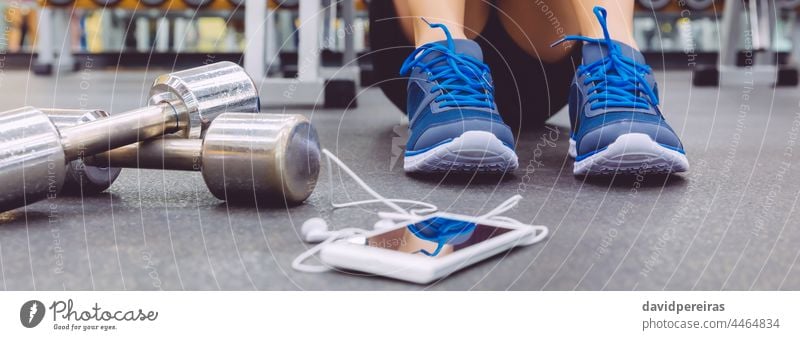 Unrecognizable sportsman sitting on the floor of the gym with dumbbells and mobile banner unrecognizable feet sneakers earphones panorama smartphone cell phone