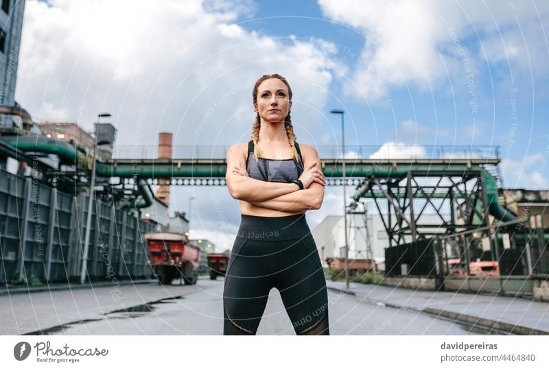 Sporty woman with crossed arms posing in front of a factory sporty self-confident looking straight proud runner clouds empowered industrial zone sportswoman