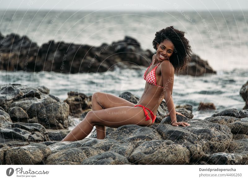 Woman sitting in rocks by the seashore woman ethnic island seaside beautiful bikini fit smile african sexy cape verde holiday horizontal happy black curly hair