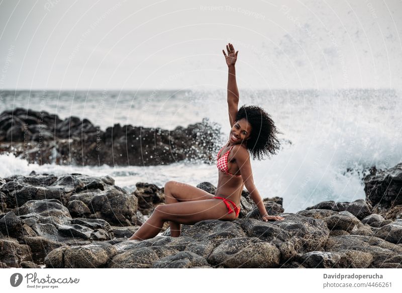 Woman sitting in rocks by the seashore woman ethnic island seaside beautiful bikini fit smile african sexy cape verde holiday horizontal happy black curly hair