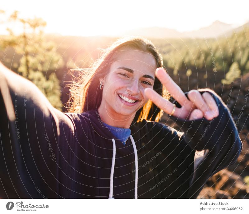 Smiling teenager showing peace gesture while taking selfie against mountain smile two fingers self portrait victory v sign cool friendly enjoy smartphone