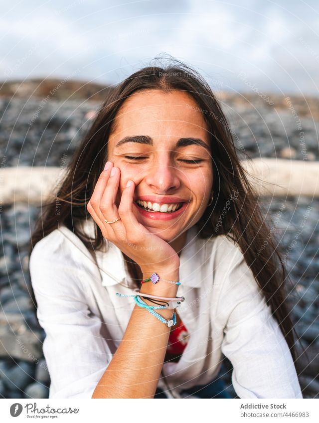 Smiling teen touching cheek on pebble shore touch cheek eyes closed mindfulness toothy smile friendly sincere beach rest portrait bracelet dreamy concentrate