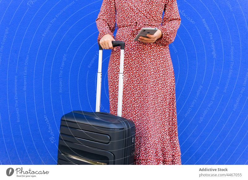 Anonymous female standing with suitcase and browsing on mobile phone against blue wall woman street luggage text message smartphone positive city red trendy