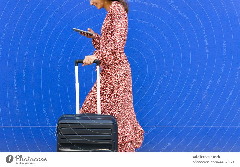Anonymous female walking with suitcase and browsing on mobile phone against blue wall woman street luggage text message smartphone positive city red trendy chat