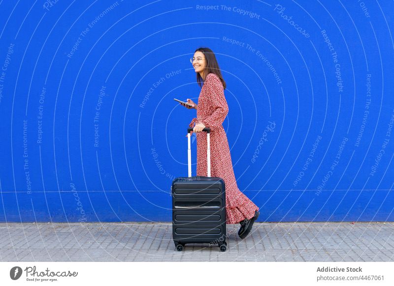 Cheerful female walking with suitcase and mobile phone against blue wall woman street smartphone luggage browsing positive city red trendy chat gadget urban
