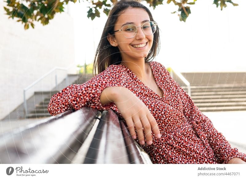 Smiling female sitting in bench in daytime woman positive style smile young cheerful wooden park rest trendy casual street lady happy toothy smile eyeglasses