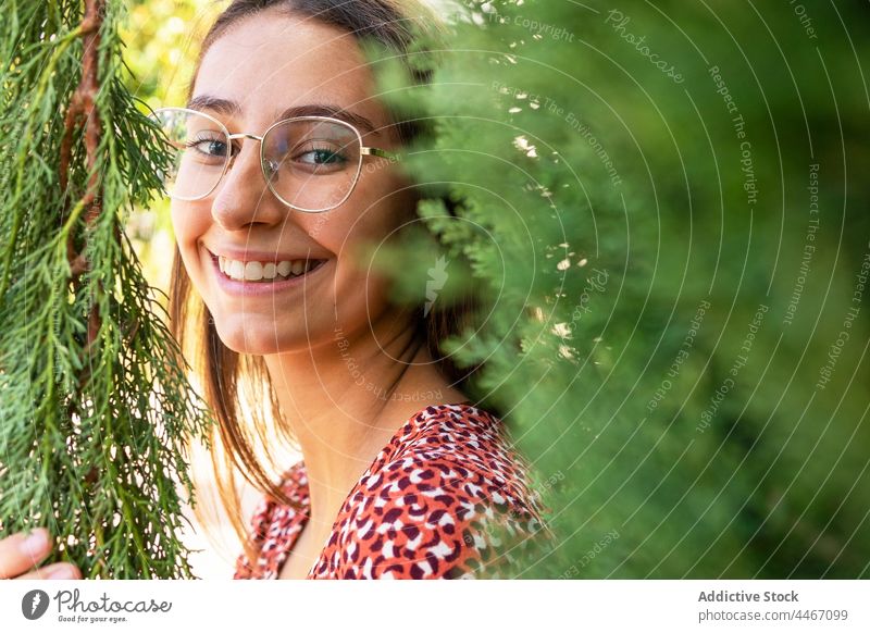 Smiling female standing among green branches woman positive cheerful smile happy toothy smile nature plant eyeglasses daylight content charming glad tree enjoy