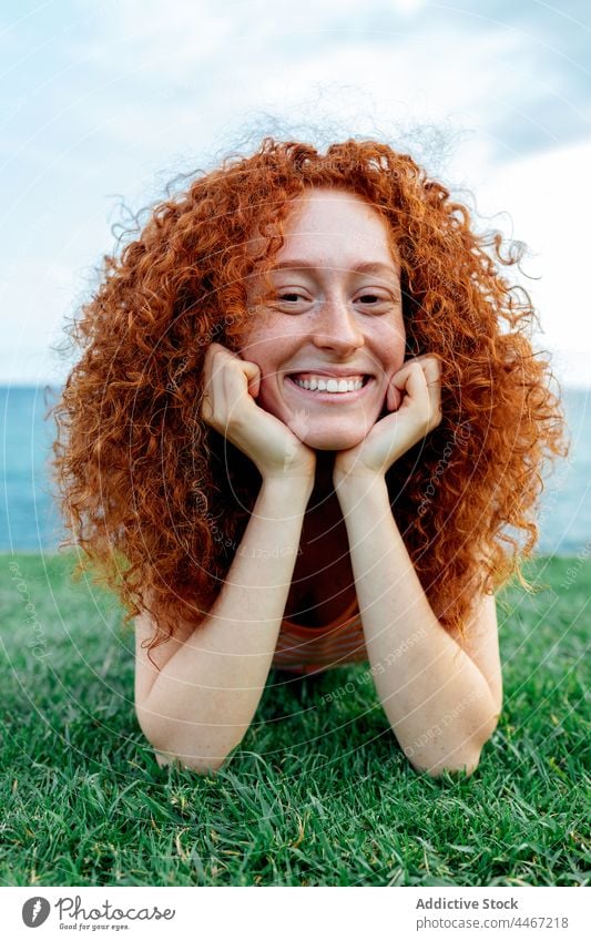 Cheerful ginger haired woman lying on grass happy carefree lawn sea relax expressive freckle female toothy smile young curly hair nature positive glad satisfied