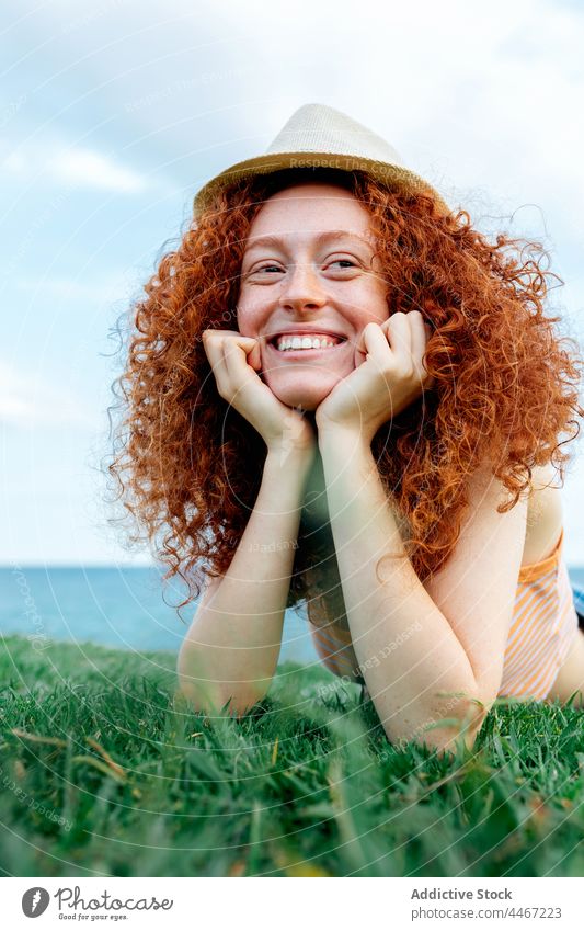 Cheerful ginger haired woman lying on grass happy carefree lawn sea relax freckle female toothy smile hat young curly hair nature positive glad satisfied water