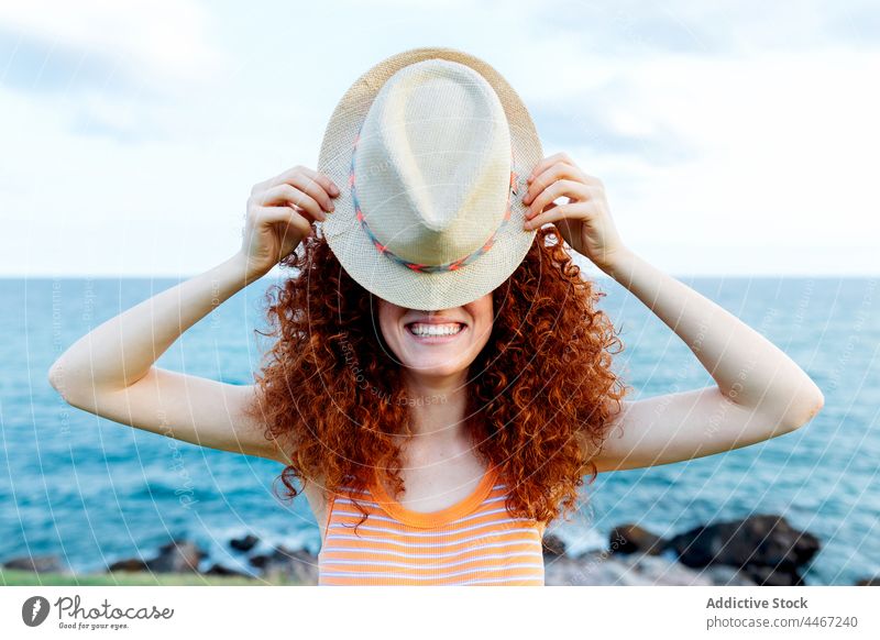 Woman covering face with hat on seashore woman hide cover face coast posture playful nature secret female curl redhead water holiday red hair vacation incognito