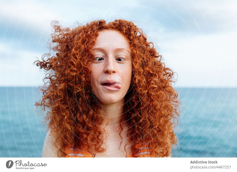 Funny woman making grimace on seashore make face playful funny show tongue behavior crazy childish female ripple cross eyes curly hair fool humor ginger hair