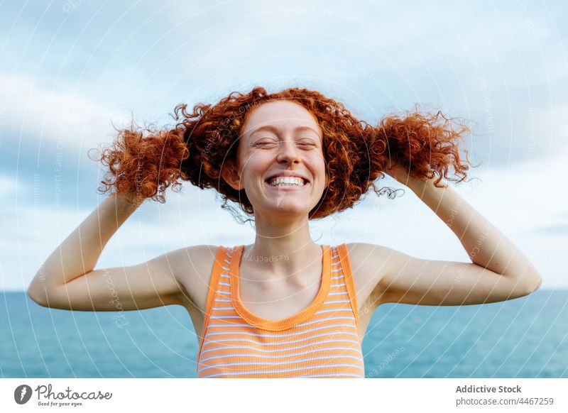 Cheerful woman touching curly hair on shore of sea funny joke personality happy expressive childish silly female redhead glee glad having fun humor portrait