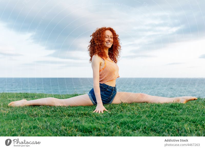 Smiling woman doing Front Split on grassy shore stretch sea front split coast posture exercise flexible yoga glad female red hair nature delight seashore water