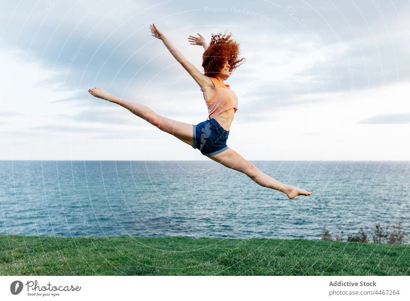 Flexible woman gracefully jumping on grassy seashore split stretch posture action flexible happy coast female freedom above ground curly hair redhead red hair