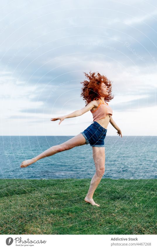 Woman gracefully dancing on grassy seashore woman dancer ballerina posture action fit flexible happy coast female freedom above ground curly hair redhead