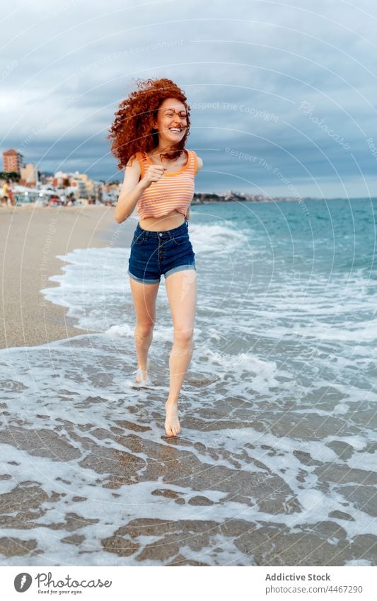 Cheerful woman running on sandy seashore beach wind happy enjoy wave foam female travel water carefree flying hair freedom coast seaside traveler redhead