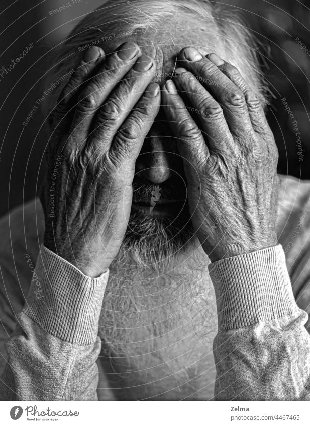 A tired, broken, resigned, preoccupied elderly man sits in front of a laptop, computer and covers his face with his hand. senior covered hands beard old older
