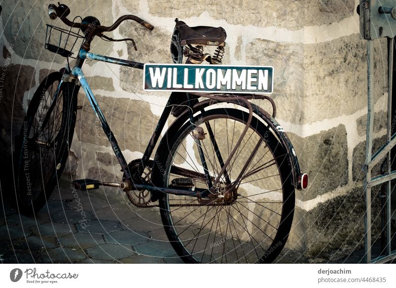 An old, dilapidated man's bicycle, leaning against a wall, with a sign in green and large white letters : WELCOME Bicycle Mobility Summer Cycling Movement