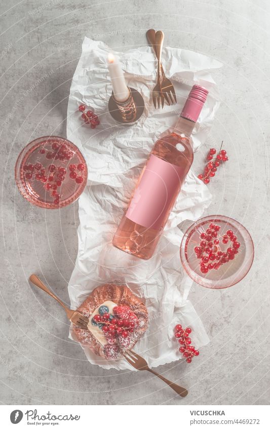 Bottle of rose wine with blank label and two wineglasses, pudding bun with red berries and forks on light background with candle. Top view bottle top view
