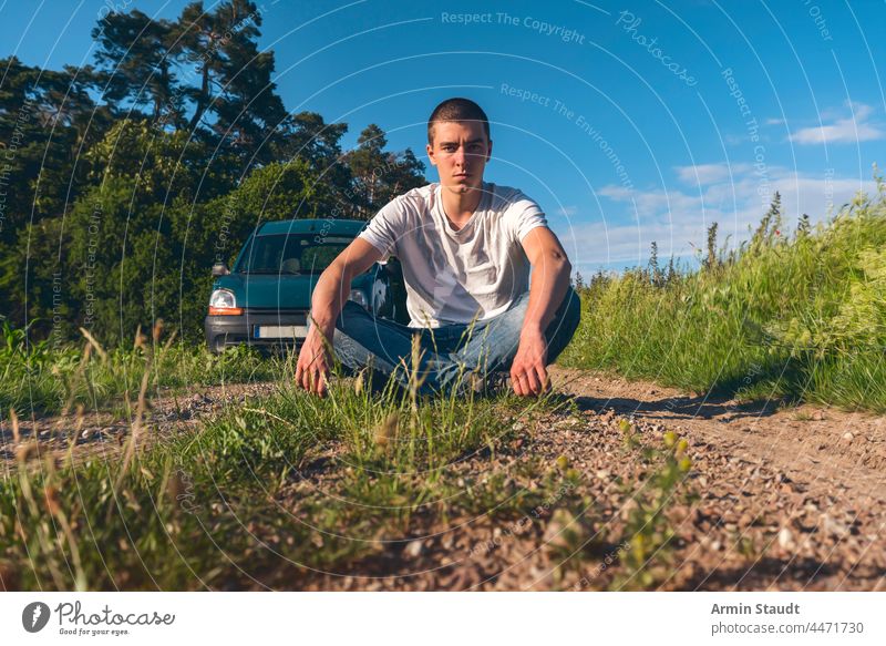 young man cross-legged on a forest path with a car in the background adventure beautiful blue casual caucasian clouds cloudscape corn enjoy far fields freedom
