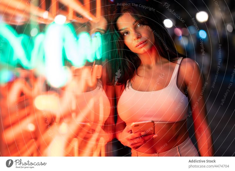 Attractive woman browsing smartphone near illuminated building glow evening street luminous online text message internet style city neon female lady charming