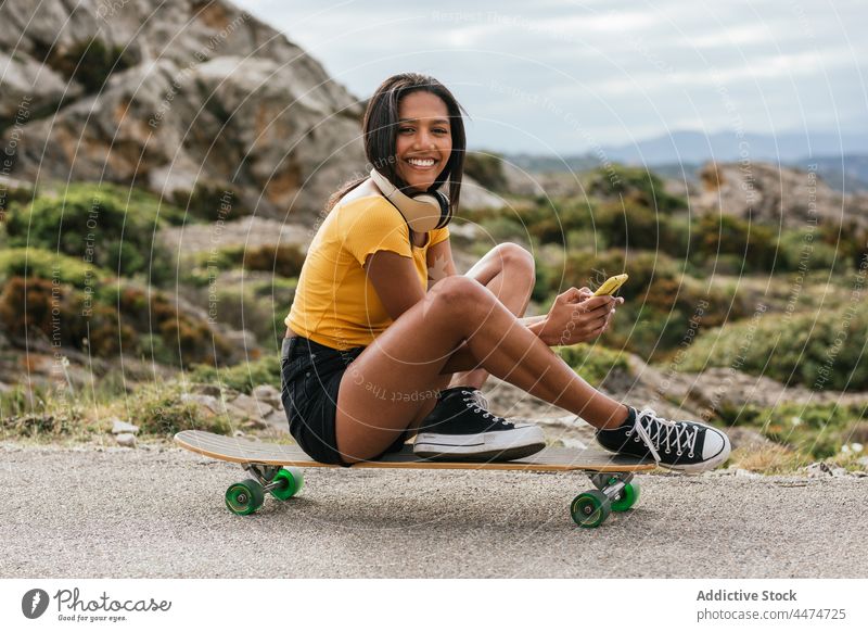 Ethnic cheerful woman using mobile phone while sitting on longboard smartphone ride watch enjoy female browsing headphones device asphalt road ethnic delight