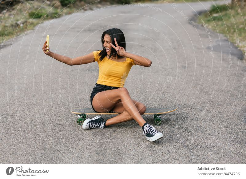 Delightful ethnic woman on longboard taking selfie on smartphone fun using peace gesture delight hobby female v sign two fingers victory happy leisure show