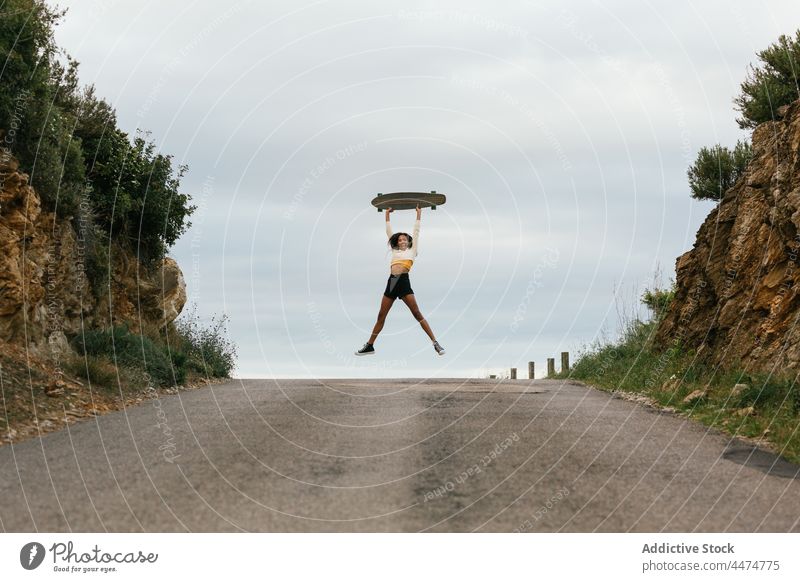 Joyful ethnic woman jumping high with longboard activity energy move joy road above ground happy female active hobby motion positive carefree excited expressive