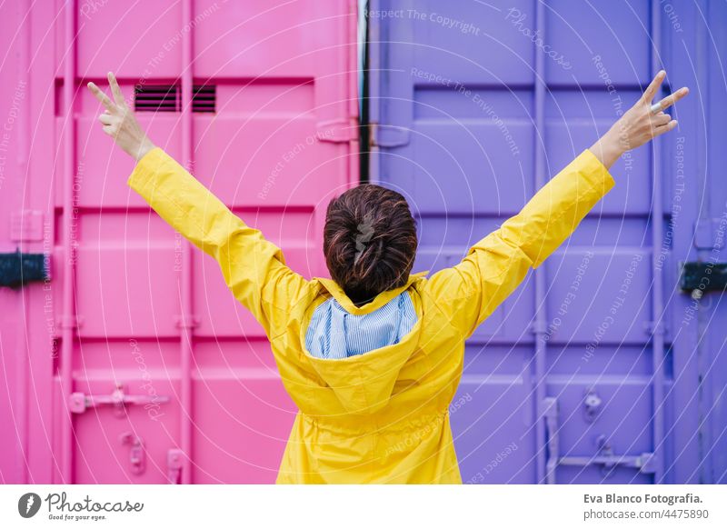 back view of woman with arms raised wearing yellow raincoat over pink and purple background. Colorful Outdoors lifestyle portrait colorful outdoors city urban