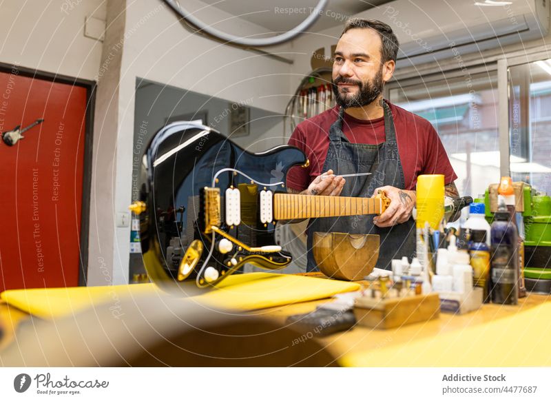 Positive man master fixing electric guitar in studio instrument skill repair equipment handicraft craftsman professional male workshop beard creative apron