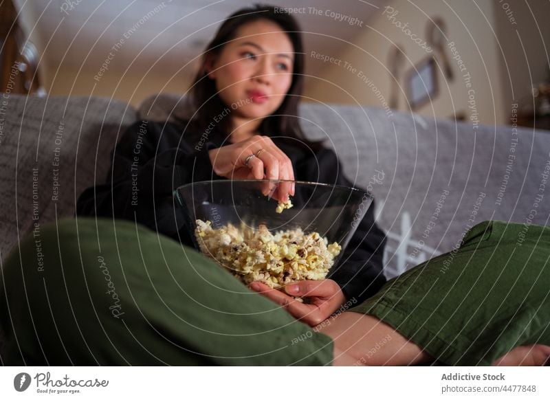 Young Asian woman sitting on sofa and eating popcorn couch legs crossed at home relax enjoy calm leisure apartment living room free time positive casual weekend