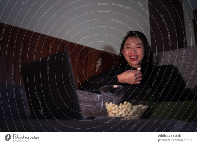 Young Asian woman sitting on sofa and eating popcorn laptop couch computer at home relax enjoy calm leisure horizontal apartment happy living room cheerful