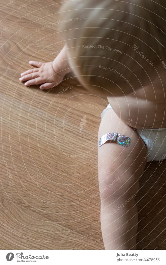 Baby looks down at metallic bandage on her leg; vaccination given to 15 month old vaccine vaccinate protect immune booster band-aid stick remove pull body belly