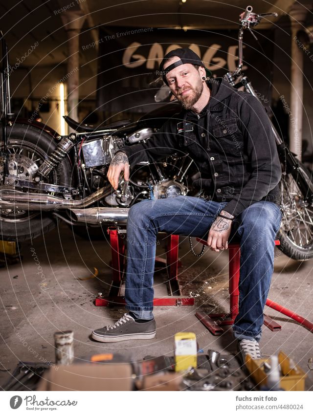 Young man sits in front of his motorcycle, biker in garage Tattooed Bikers Motorcycle Chopper Vehicle Facial hair cappy Frock Screwdriver Pride Man Transport