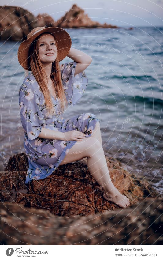 Stylish woman sitting on seashore evening beach coast summer rock dress hat sunset female charming nature water young style stone dreamy calm attractive sundown