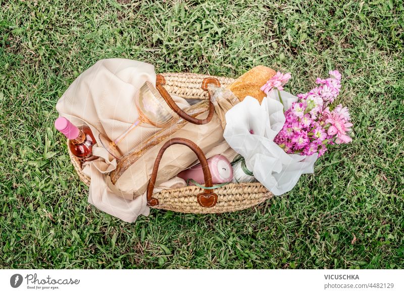 Picnic basket with wine, baguette, wine glass, flowers, candle and picnic blanket on green grass background. Romantic picnic preparation in summer. Top view.