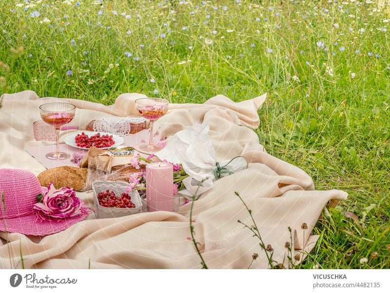 Picnic blanket with wine glasses, rose wine, candles, sun hat, fresh berries and baguette on green grass. Romantic brunch outdoor with food and drinks in summer. Front view.
