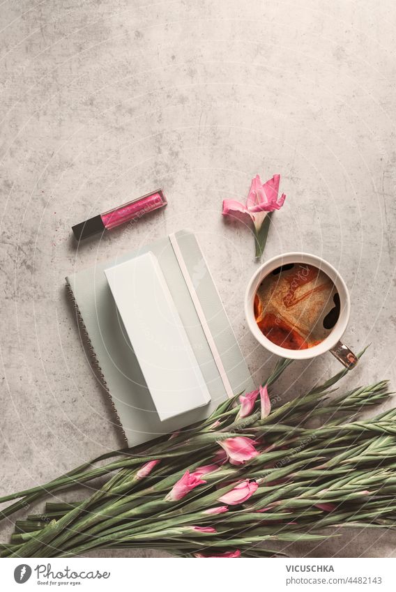 Coffee, notebook, pink flowers and lip gloss on pale grey concrete background. Feminine morning routine. Top view with copy space. coffee feminine top view