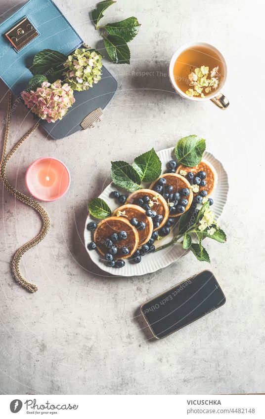 Feminine breakfast lifestyle with blueberries pancakes, cup of tea , burning candle and smartphone with black screen, blue handbag with hydrangea on light grey concrete table. Top view.