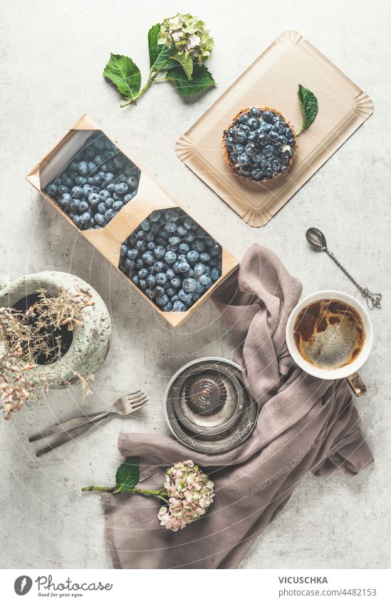 Flat lay with blueberries cake, coffee, wooden basket with fresh blueberries,dishcloth, water glass and flowers on white background. Cozy tea time with homemade pastry and hot drinks. Top view.